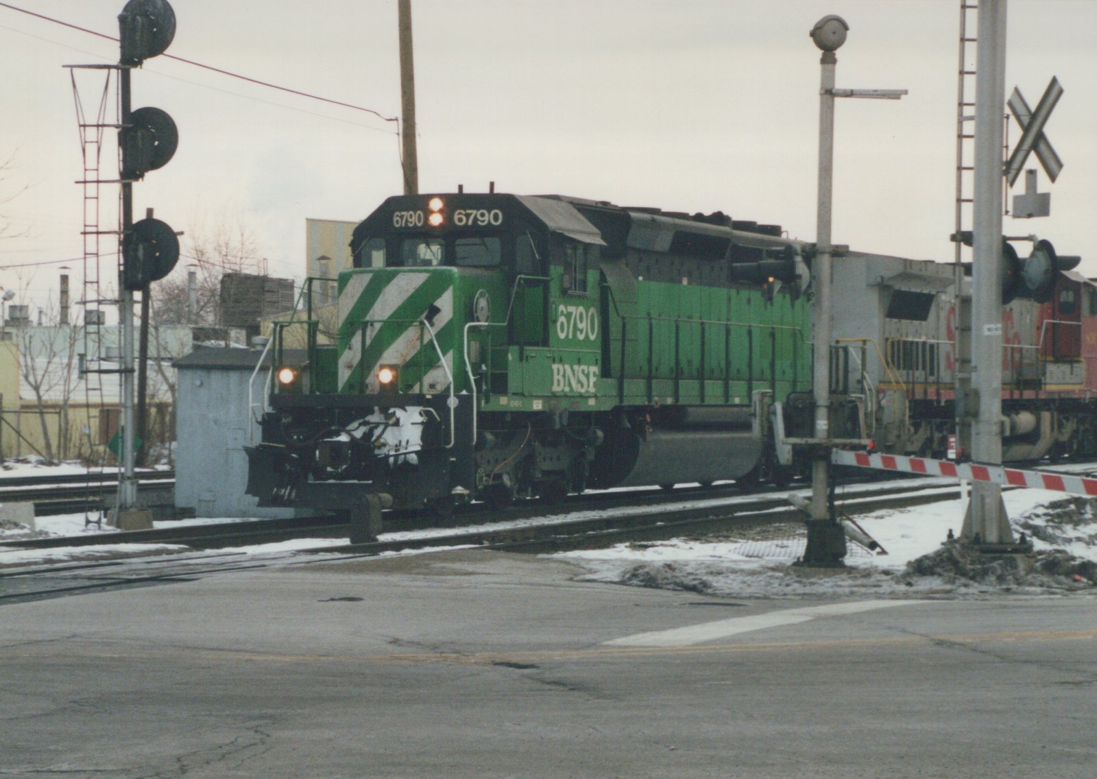 A green BNSF locomotive numbered 6790 is stopped at a railroad crossing with snow on the ground and a cloudy sky.