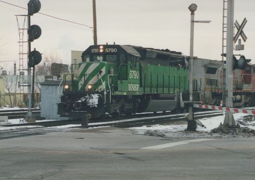A green BNSF locomotive numbered 6790 is stopped at a railroad crossing with snow on the ground and a cloudy sky.