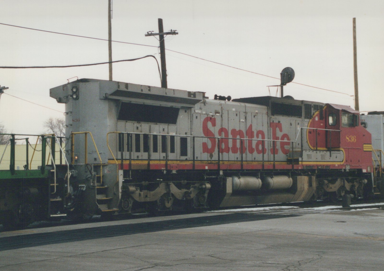 A side view of a Santa Fe SD45 diesel locomotive, number 836, in a silver, red, and yellow livery, stopped on tracks in a yard with snow on the ground.