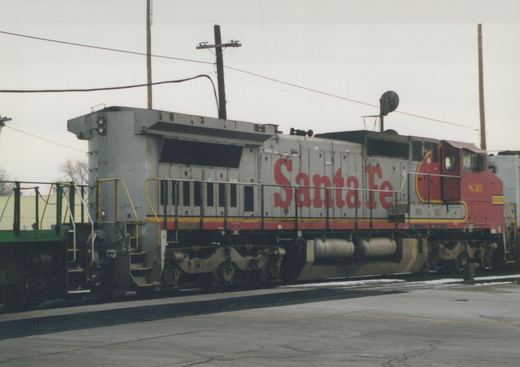 A side view of a Santa Fe SD45 diesel locomotive, number 836, in a silver, red, and yellow livery, stopped on tracks in a yard with snow on the ground.