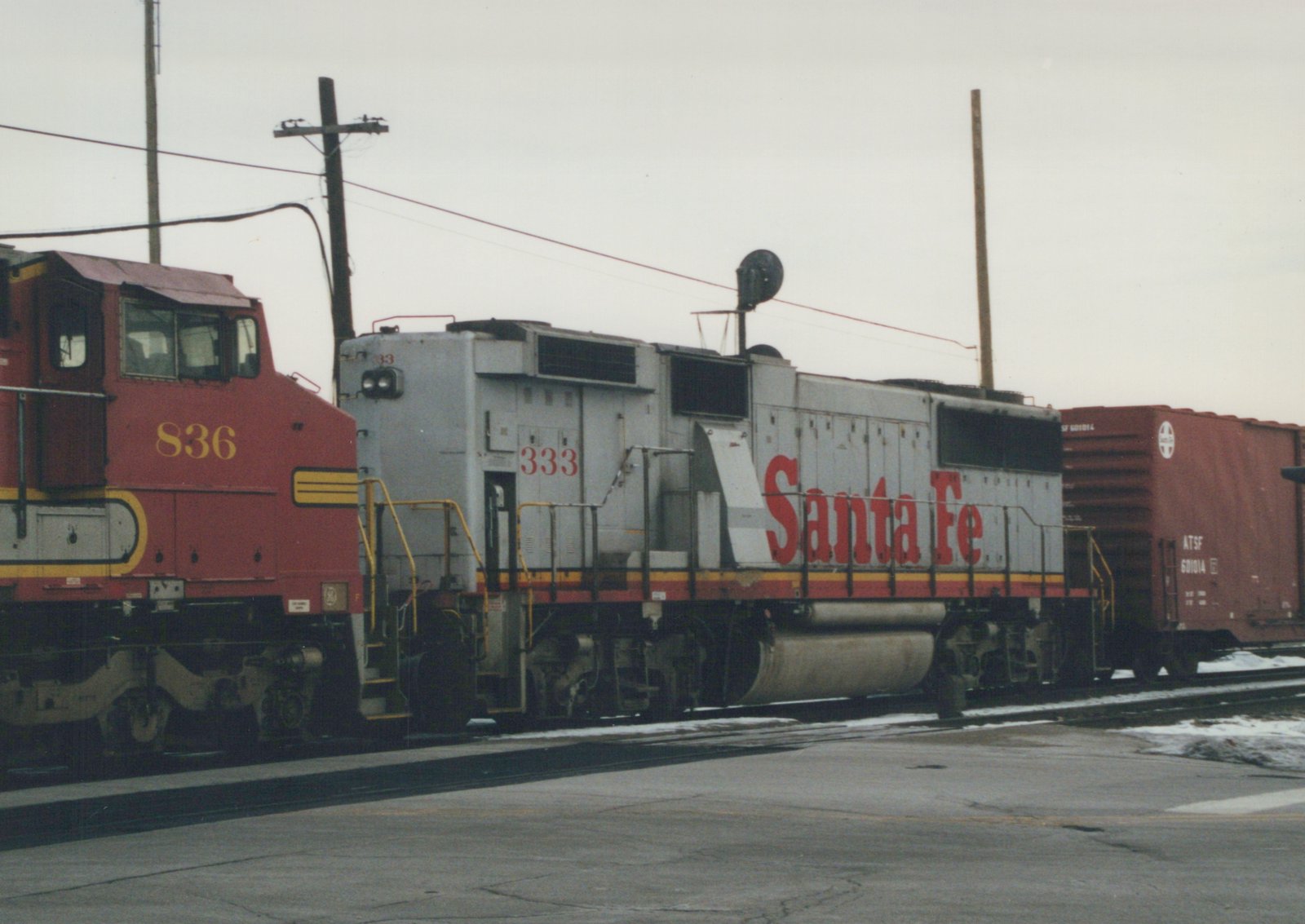 Two diesel locomotives, one gray with red lettering and the other red with yellow stripes, sit coupled to a red freight car on railroad tracks with snow on the ground.