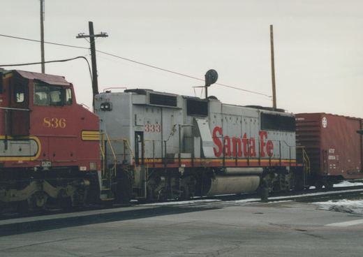 Two diesel locomotives, one gray with red lettering and the other red with yellow stripes, sit coupled to a red freight car on railroad tracks with snow on the ground.