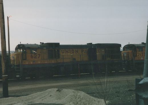 Two orange and black Union Pacific diesel locomotives, numbers 627 and 2652, are parked in a gravel rail yard with a utility pole and bare shrubs in the foreground under a grey sky.