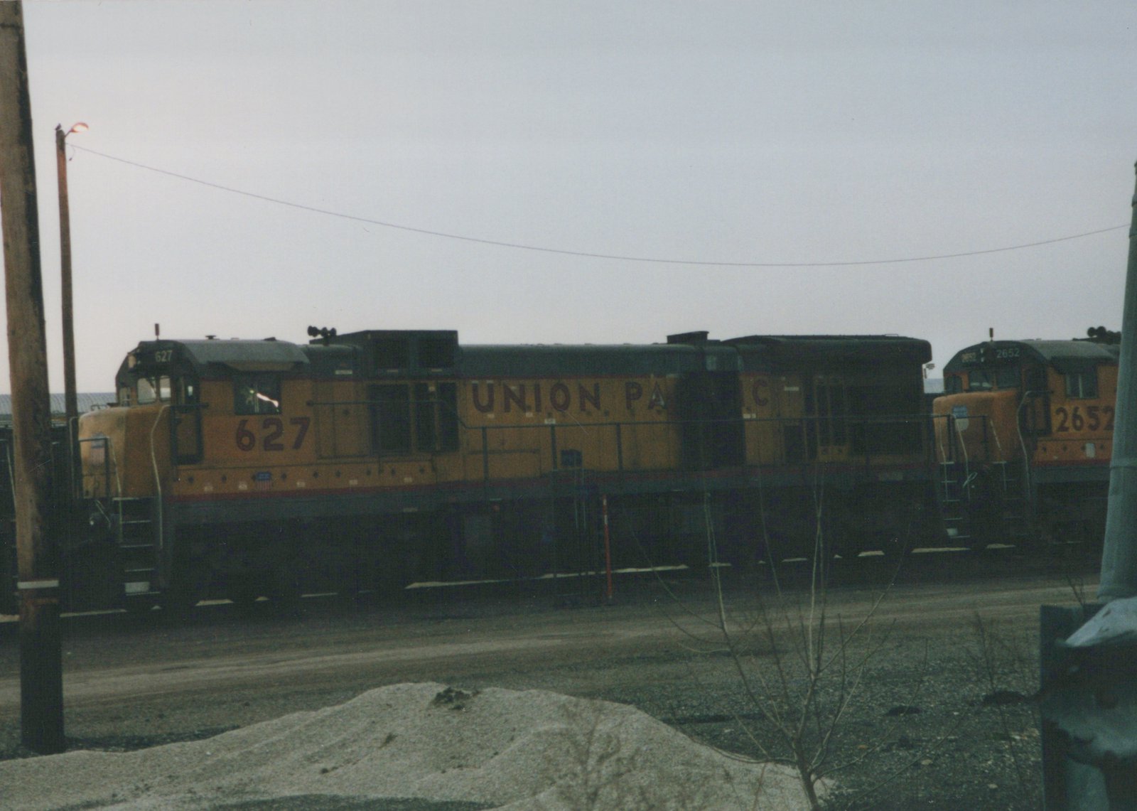 Two orange and black Union Pacific diesel locomotives, numbers 627 and 2652, are parked in a gravel rail yard with a utility pole and bare shrubs in the foreground under a grey sky.