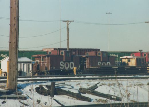 A dark brown Soo Line caboose with the number 141 is parked in a snow-covered rail yard, with green and red boxcars visible in the background.