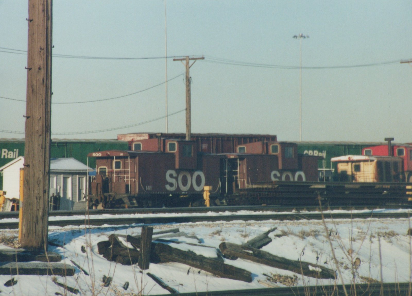 A dark brown Soo Line caboose with the number 141 is parked in a snow-covered rail yard, with green and red boxcars visible in the background.