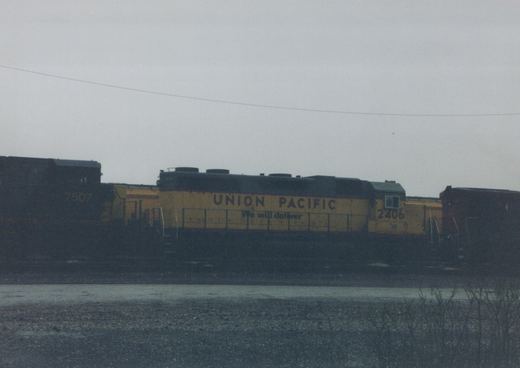 A Union Pacific diesel locomotive, number 2406, in yellow and black, sits on tracks in a rail yard under a grey sky.