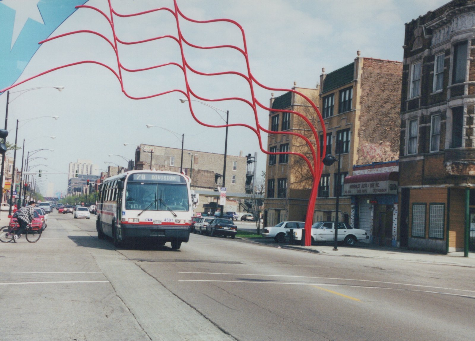 A white and red Chicago Transit Authority bus, number 4591, drives down a city street with a large red abstract sculpture overhead and brick buildings lining the road.