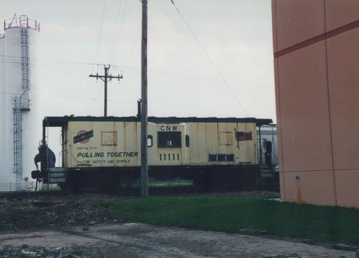 A cream-colored Chicago and North Western caboose numbered 11111 with a green roof and 'Pulling Together' slogan, parked beside a grassy track with a utility pole and silo in the background under an overcast sky.