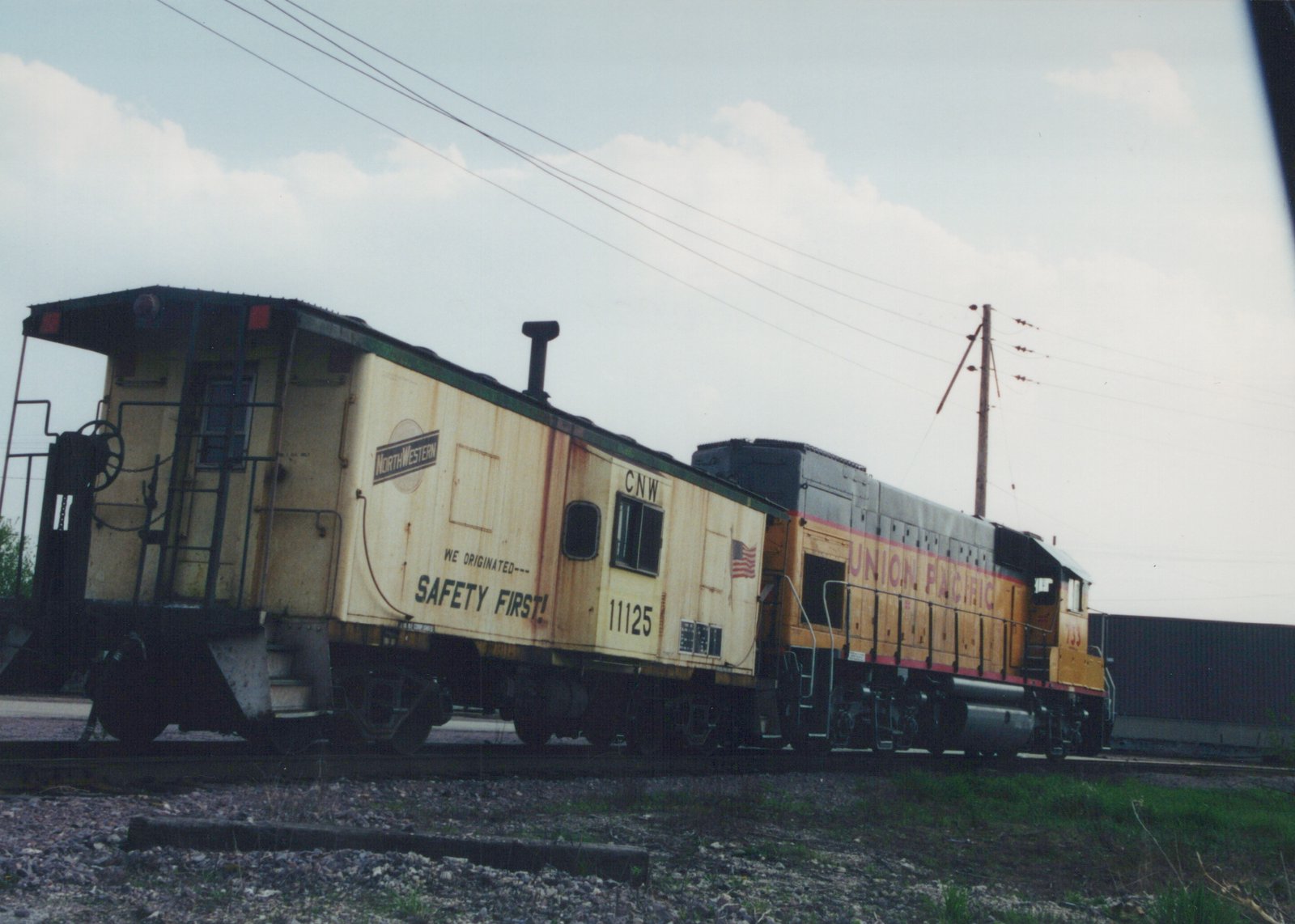 A yellow and gray Union Pacific locomotive pulls a cream-colored Chicago & North Western caboose along a railroad track on a cloudy day, with utility poles in the background.