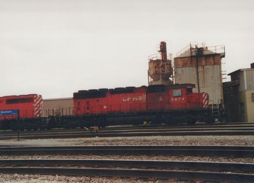 A red Canadian Pacific locomotive, number 5674, sits on tracks in a rail yard with other tracks and industrial buildings visible.