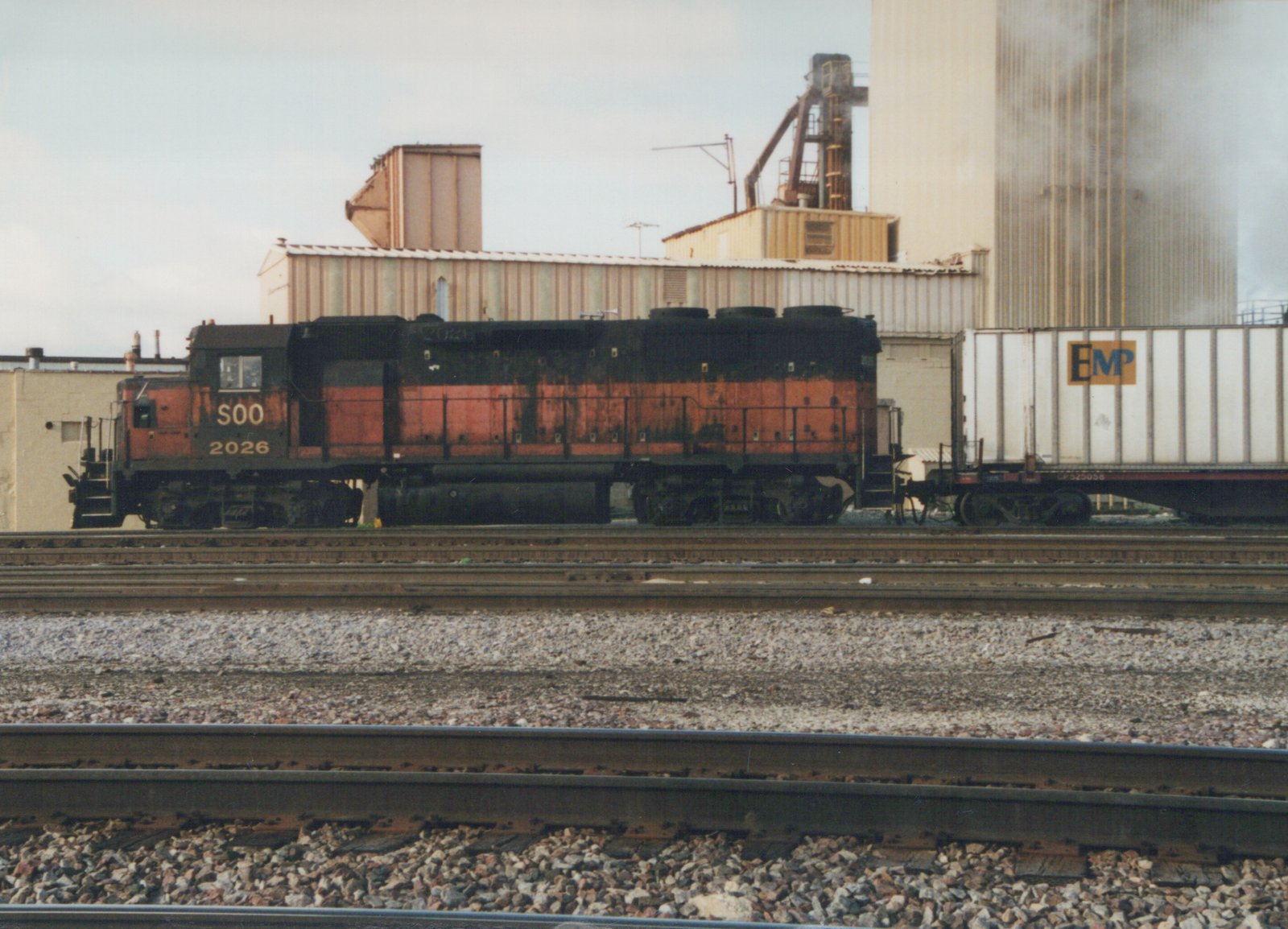 A side view of a black and orange Soo Line diesel locomotive, number 2026, pulling a white freight car with an EMP logo, on a set of railroad tracks.