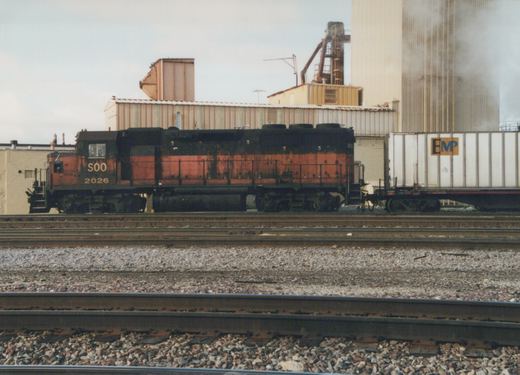 A side view of a black and orange Soo Line diesel locomotive, number 2026, pulling a white freight car with an EMP logo, on a set of railroad tracks.