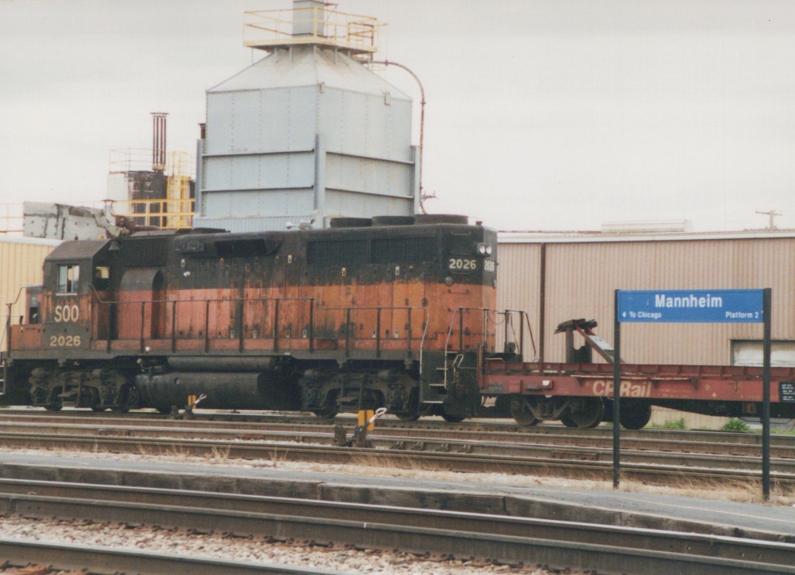 A side view of a SOO Line diesel locomotive, number 2026, stopped at a station platform next to a CP Rail flatcar, with a blue sign reading 'Mannheim' in the background.
