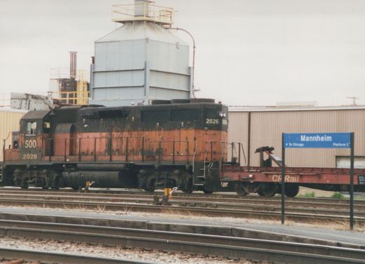 A side view of a SOO Line diesel locomotive, number 2026, stopped at a station platform next to a CP Rail flatcar, with a blue sign reading 'Mannheim' in the background.
