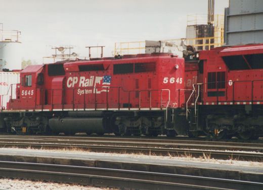 A bright red CP Rail System diesel locomotive, number 5645, is shown from the side in a rail yard under an overcast sky, with industrial structures in the background.