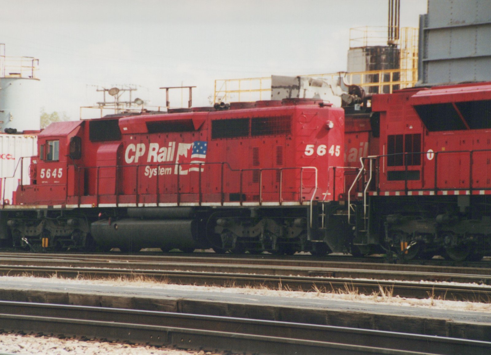 A bright red CP Rail System diesel locomotive, number 5645, is shown from the side in a rail yard under an overcast sky, with industrial structures in the background.