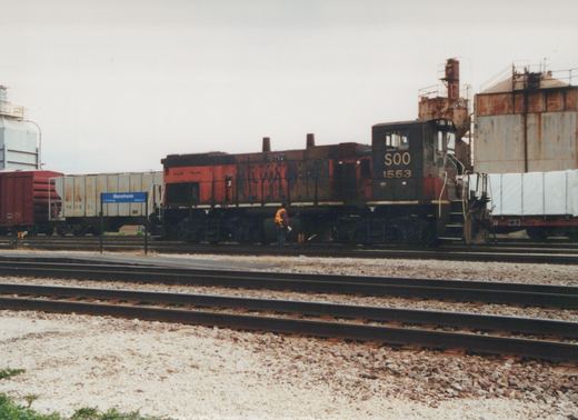 A black and red Soo Line locomotive numbered 1553 stands on tracks at Mannheim station, with a worker in high-visibility vest nearby and several freight cars, including a white boxcar and a red boxcar, in the background under an overcast sky.