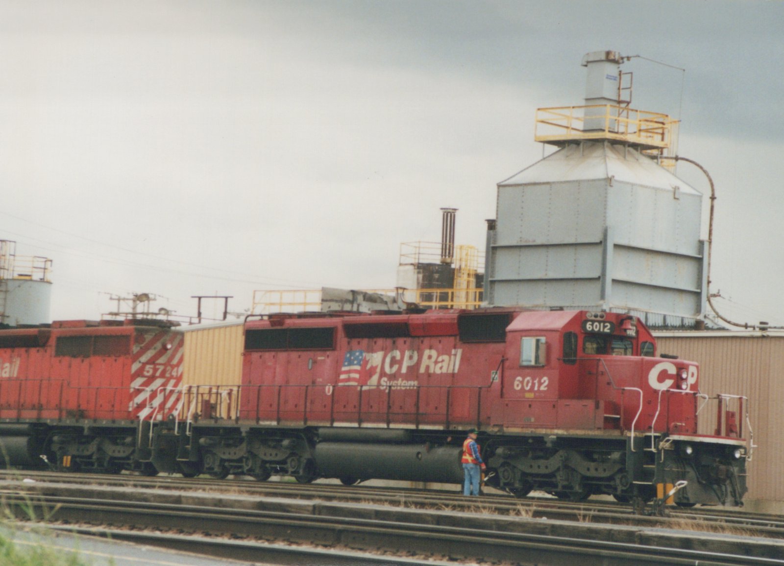 A red CP Rail System diesel locomotive numbered 6012 is stopped on the tracks in a rail yard, with a second locomotive behind it and industrial buildings in the background.