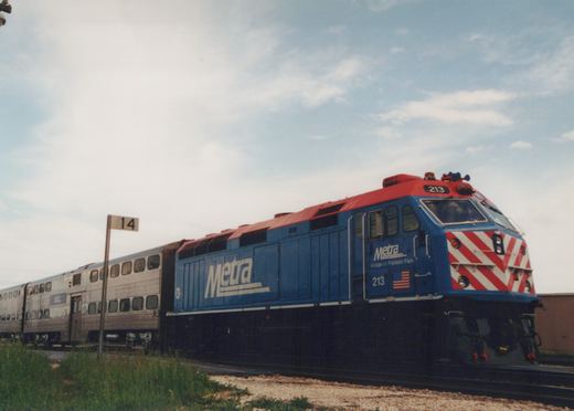 A blue and red Metra locomotive numbered 213 pulls silver double-decker gallery cars on a sunny day in a rail yard.