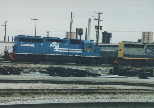 Side view of a blue Conrail locomotive numbered 8869 next to a gray CSX locomotive numbered 8247, both on parallel tracks in a gravel yard with industrial buildings in the background.