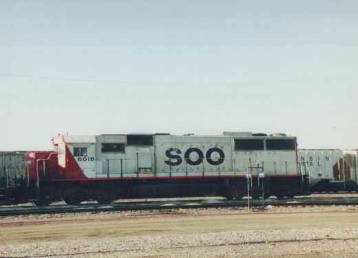 A side view of a SOO Line diesel locomotive, number 6018, painted white with a red nose and stripe, standing on tracks in a gravel yard under a clear sky.
