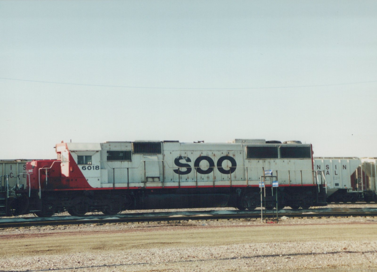 A side view of a SOO Line diesel locomotive, number 6018, painted white with a red nose and stripe, standing on tracks in a gravel yard under a clear sky.