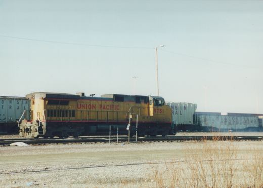 A yellow and gray Union Pacific diesel locomotive numbered 9751 is shown in profile, pulling a line of gray Wisconsin Central freight cars on a clear winter day.