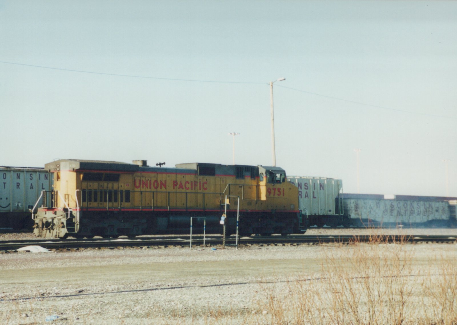 A yellow and gray Union Pacific diesel locomotive numbered 9751 is shown in profile, pulling a line of gray Wisconsin Central freight cars on a clear winter day.