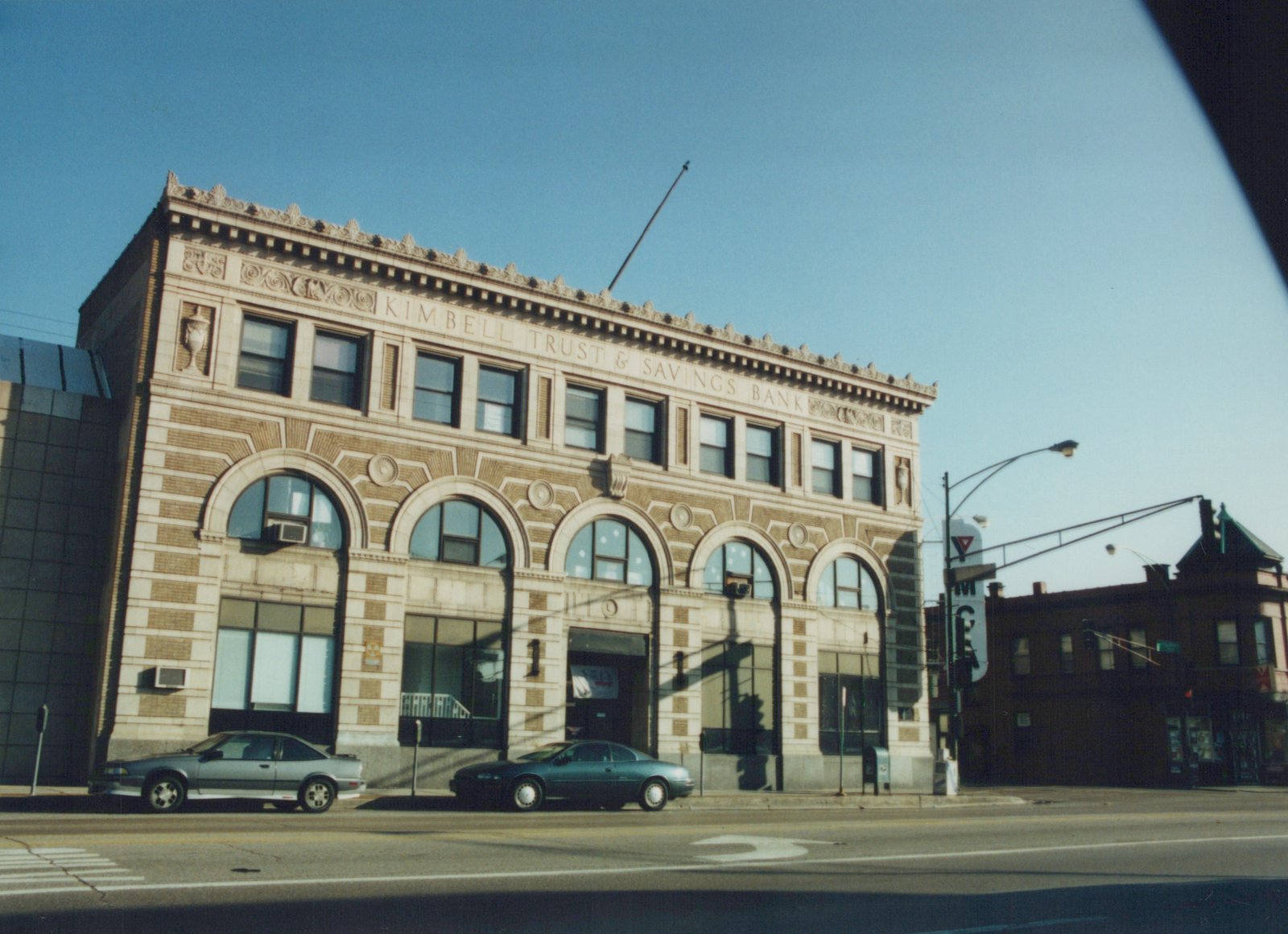 A historic two-story bank building with arched windows and stone facade, labeled 'KIMBELL TRUST & SAVINGS BANK', with two parked cars on a street under a clear blue sky.