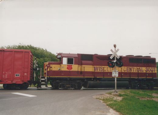 A maroon and yellow Wisconsin Central Ltd diesel locomotive numbered 3007 is stopped at a railroad crossing with its red lights flashing.