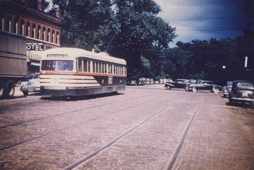 A white, orange, and blue streetcar with the number 1023 on its front is stopped at an intersection on a cobblestone street, with vintage cars and a brick building in the background.