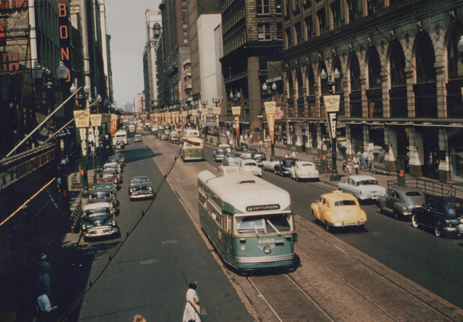 A green and cream PCC streetcar numbered 4263 travels down a busy Chicago street with vintage cars and pedestrians, under clear skies.