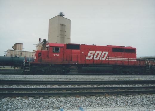Side view of a red Soo Line diesel locomotive numbered 761, coupled to a black tank car on a gravel-ballasted railroad yard.