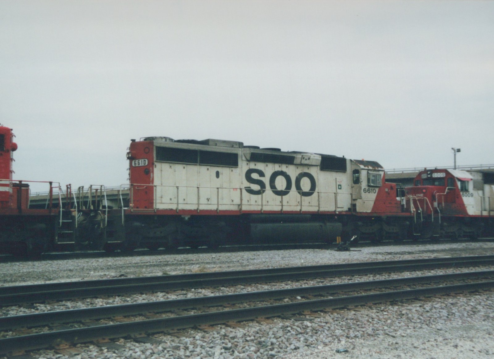 Side view of two Soo Line diesel locomotives, numbers 6610 and 6009, in white and red livery, parked on parallel tracks in a gravel yard under a grey sky.