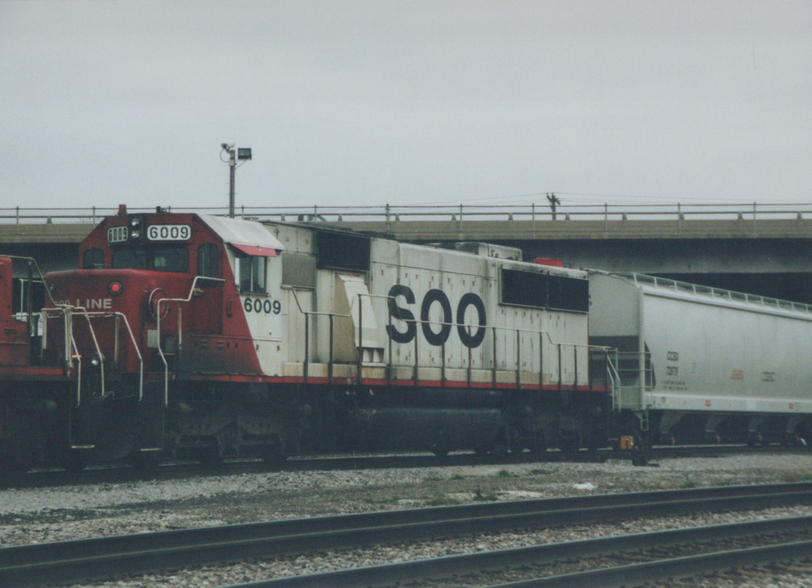A red and white Soo Line locomotive numbered 6009 pulls a white freight car under a concrete bridge on an overcast day.