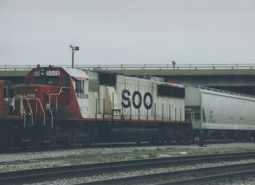 A red and white Soo Line locomotive numbered 6009 pulls a white freight car under a concrete bridge on an overcast day.