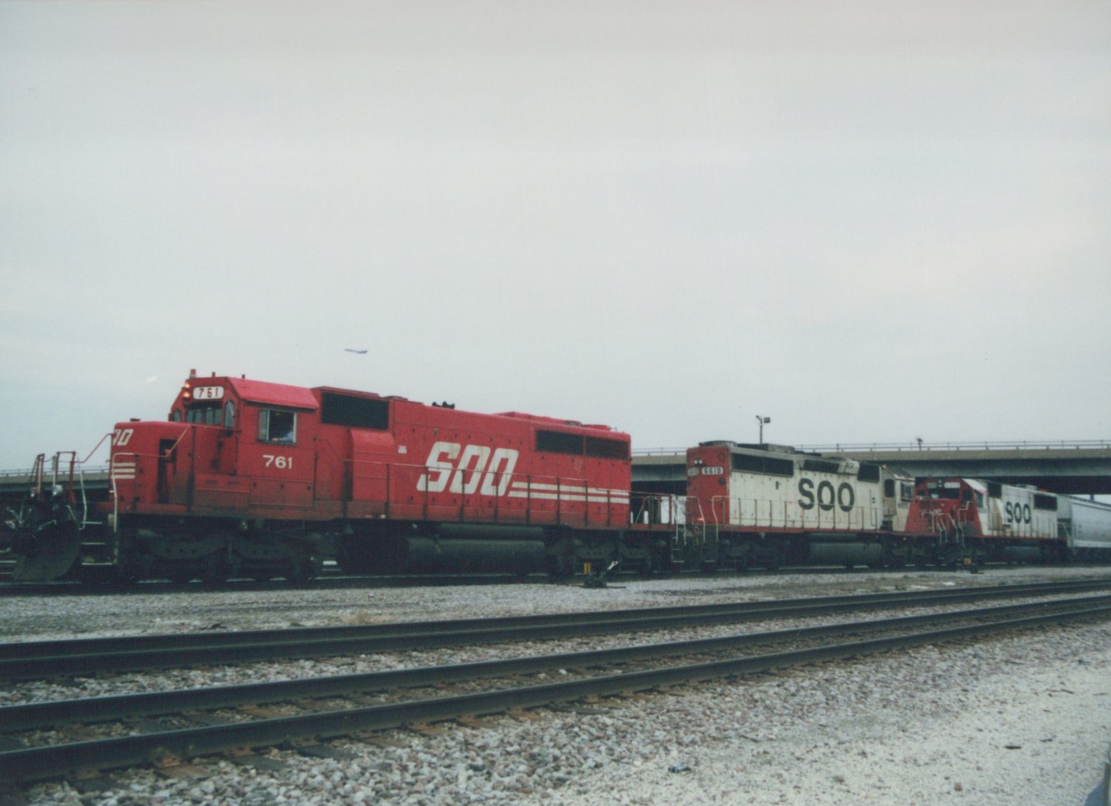 A side view of three red and white SOO Line diesel locomotives parked on railroad tracks with an elevated highway overpass in the background under an overcast sky.