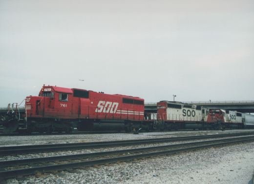 A side view of three red and white SOO Line diesel locomotives parked on railroad tracks with an elevated highway overpass in the background under an overcast sky.