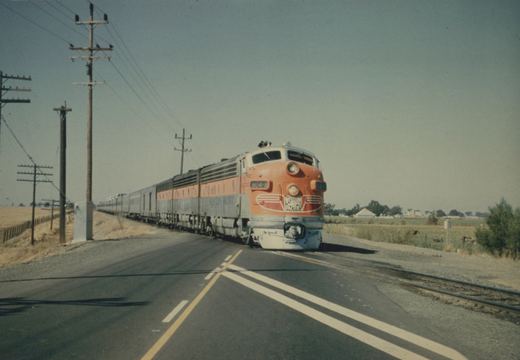 A Western Pacific FP7A diesel locomotive, number 804-A, in orange and gray livery, pulls a line of matching passenger cars across a paved road in a rural, dry field setting under a clear sky.