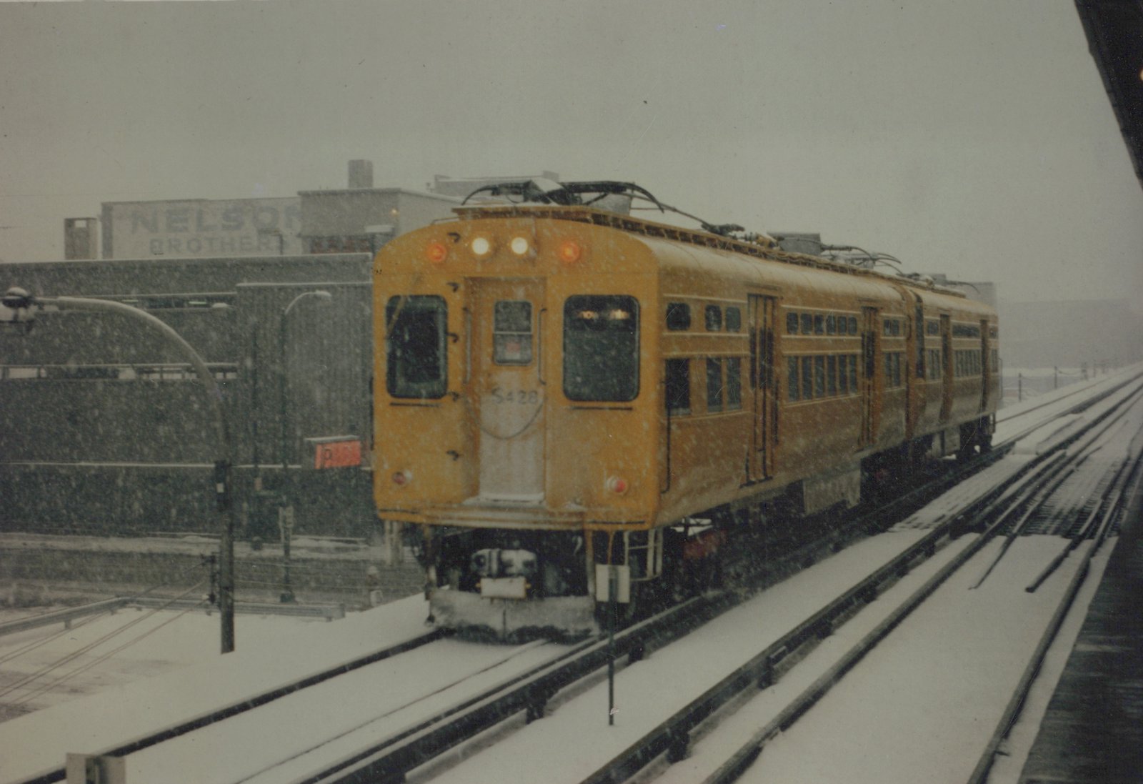 A yellow interurban train with the number S428 is moving on a snow-covered elevated track, with the words 'NELSON BROTHERS' visible on a building in the background.