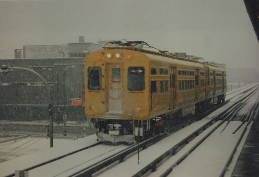 A yellow interurban train with the number S428 is moving on a snow-covered elevated track, with the words 'NELSON BROTHERS' visible on a building in the background.