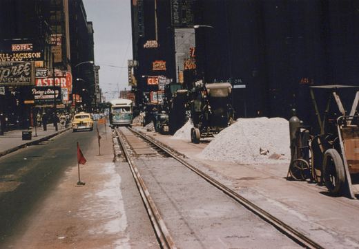 A vintage street scene in Chicago showing a green and cream PCC streetcar on tracks, with GMC buses and a yellow car nearby. Construction equipment and piles of gravel line the street, while neon signs for hotels and restaurants adorn the tall buildings.