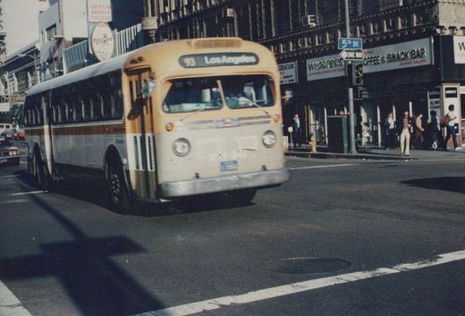 A yellow and cream city bus with the destination '93 Los Angeles' is turning a corner at an intersection marked '5th St' and 'ONE WAY'. Pedestrians are crossing the street and a 'WHOOPEE'S' snack bar is visible on the corner.