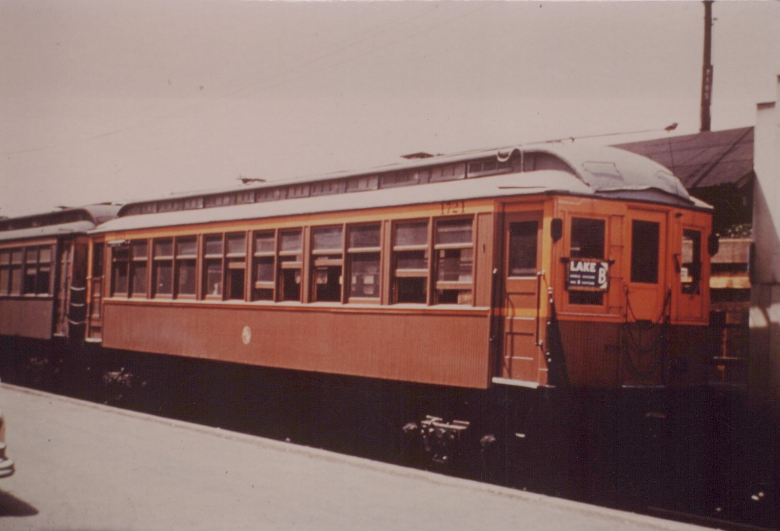 A side and front view of a vintage orange and maroon interurban streetcar with the number 1721 and a destination sign reading 'LAKE B' on a city street with overhead wires.