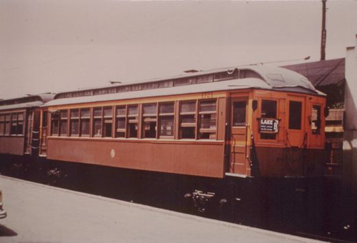 A side and front view of a vintage orange and maroon interurban streetcar with the number 1721 and a destination sign reading 'LAKE B' on a city street with overhead wires.