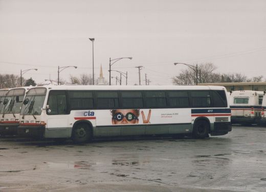 A white Chicago Transit Authority motor bus, number 4717, is parked side-on in a wet paved yard. Other buses are visible in the background. The side of the bus has a large ad with a person's face and the text 'jump in january 15 2000'.