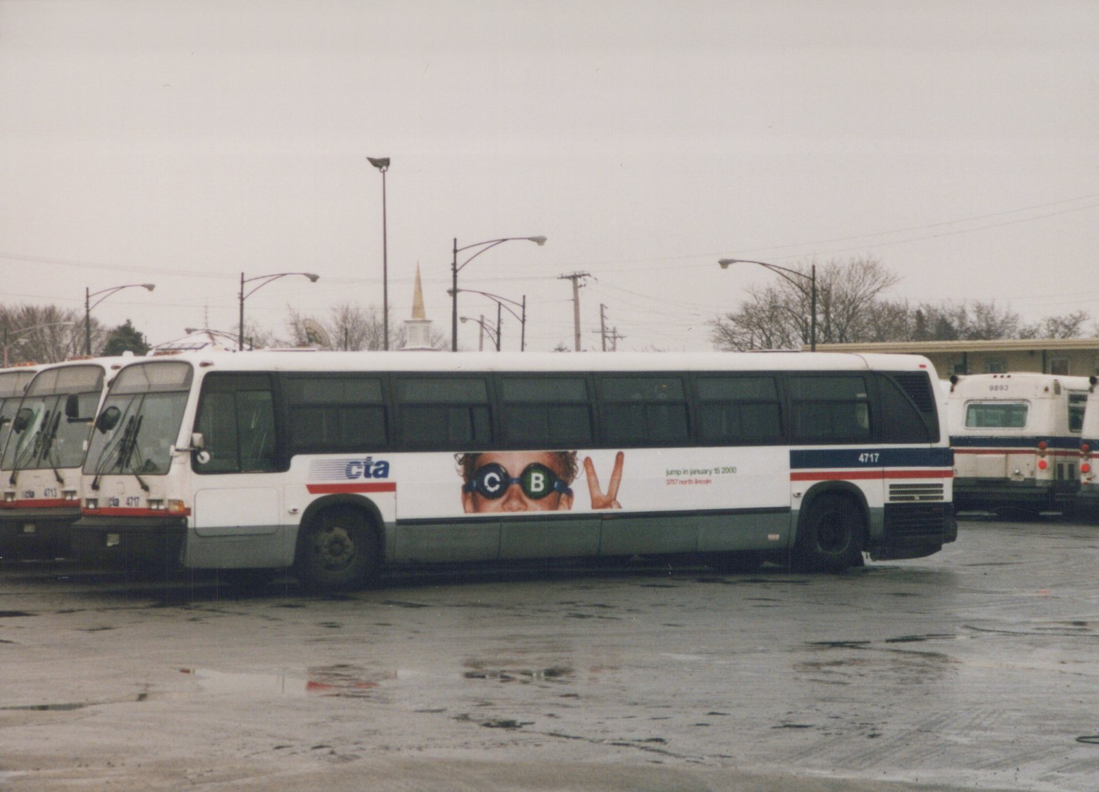 A white Chicago Transit Authority motor bus, number 4717, is parked side-on in a wet paved yard. Other buses are visible in the background. The side of the bus has a large ad with a person's face and the text 'jump in january 15 2000'.