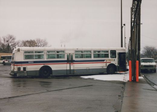Side view of a white, blue and red Chicago Transit Authority bus, fleet number 9892, parked in a paved yard with patches of snow and another bus in the background under an overcast sky.