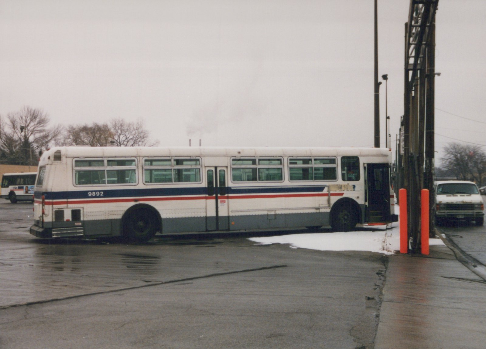 Side view of a white, blue and red Chicago Transit Authority bus, fleet number 9892, parked in a paved yard with patches of snow and another bus in the background under an overcast sky.